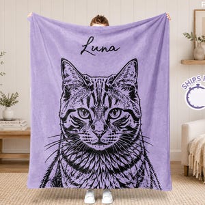Op de afbeelding: Een lila deken met een zwart-wit illustratie van een kattengezicht. De naam van de kat, "Luna", staat in zwart schrift boven de afbeelding. De deken wordt door een persoon vastgehouden.