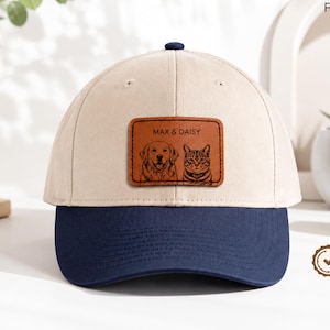 May include: A two-tone baseball cap featuring a beige crown and a dark blue brim. Centered on the front is a rectangular brown leather patch engraved with a line drawing of a dog and a cat, with the text MAX & DAISY printed above the animals.