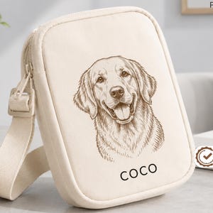 May include: A cream-colored rectangular crossbody bag with a zipper closure and an adjustable strap. The front features a brown line-art illustration of a golden retriever dog and the name COCO printed in black capital letters below the dog.