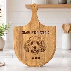 May include: A light brown wooden pizza peel serving board featuring a laser-engraved illustration of a dog face. The text reads CHARLIE'S PIZZERIA and EST. 2026. The board has a long handle with a hole at the top for hanging and a rounded base.