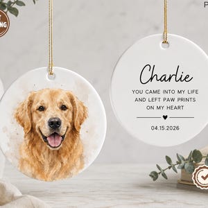 Peut inclure: Deux ornements ronds en céramique blanche avec des cordons dorés. L'un présente une illustration à l'aquarelle d'un golden retriever. L'autre affiche le nom Charlie, le texte YOU CAME INTO MY LIFE AND LEFT PAW PRINTS ON MY HEART et 04.15.2026.