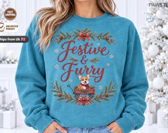 May include: A teal blue crewneck sweatshirt featuring a graphic print with the text Festive & Furry in red script. The design includes a corgi wearing a red scarf inside a red wagon, surrounded by a wreath of pine branches, pinecones, and bows.