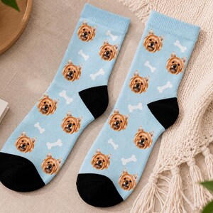May include: A pair of light blue socks featuring a repeating pattern of a dog's face and white bone shapes. The socks have black heels and toes. The dog's face is brown and tan.