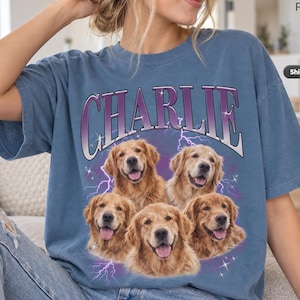 May include: A blue short-sleeve t-shirt featuring a graphic print of five golden retriever dogs. Above the dogs, the name CHARLIE is printed in large purple and white letters. The background of the graphic includes purple lightning and stars.