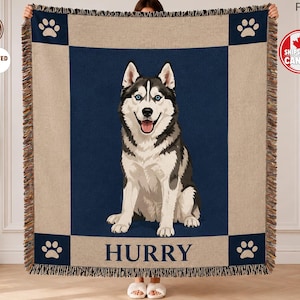 Peut inclure: Un plaid tissé présentant un panneau central bleu foncé avec une illustration de Husky sibérien noir et blanc. La bordure est beige avec des carrés d'angle bleu foncé contenant des empreintes de pattes blanches. Le nom HURRY est imprimé en bleu.