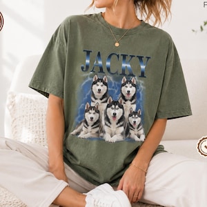 Puede incluir: Una camiseta de manga corta color verde oliva con un estampado gráfico de cinco perros Husky siberianos agrupados. Sobre los perros, el nombre JACKY está impreso en una fuente azul. El diseño incluye un fondo azul con aspecto de nubes.