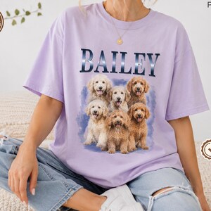 May include: A light purple short-sleeve t-shirt featuring the name BAILEY in dark blue lettering above a graphic of six golden-colored doodle dogs. The dogs are arranged in a group against a soft blue watercolor-style background.