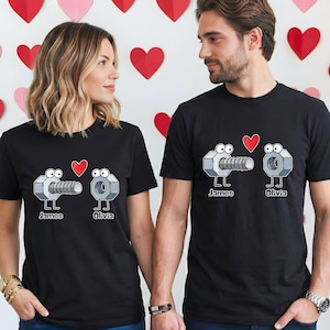 May include: Black t-shirts featuring a cartoon design of a bolt and a nut with heart accents. The names "James" and "Olivia" are printed below the characters. The background includes pink and red heart decorations.