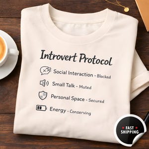 Może przedstawiać: Kremowa koszulka z napisem "Introvert Protocol" oraz powiązanymi ikonami i frazami. Frazy obejmują "Social Interaction - Blocked", "Small Talk - Muted", "Personal Space - Secured" i "Energy - Conserving."