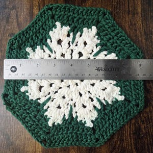 May include: A handmade, crocheted doily in a snowflake design. The doily is primarily dark green with a white snowflake pattern in the center. The doily measures approximately 7 inches in diameter.