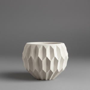 Peut inclure: Un pot de fleurs en béton blanc avec un extérieur géométrique à facettes présentant un motif vertical en losanges répété. Le récipient possède une large ouverture circulaire sur le dessus et une base arrondie, avec une finition mate texturée.