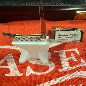Può includere: Un piccolo coltello tascabile aperto, con un manico a pois blu e bianchi, appoggiato su un supporto bianco a forma di incudine. La lama del coltello è aperta e una piccola scatola con il testo "Item No 08825" è accanto.