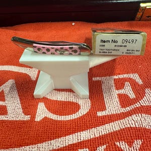 Può includere: Un piccolo coltello tascabile con un motivo rosa e marrone a pois e una lama argentata, appoggiato su un supporto bianco a forma di incudine. Il coltello è accanto a una scatola con il testo "Item No 09497".
