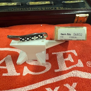 Può includere: Un piccolo coltello tascabile chiuso con un manico a pois bianchi e neri poggia su un supporto a forma di incudine bianca. Una piccola scatola con la scritta "Item No 06832" è accanto al coltello. Una vetrina con altri coltelli è sullo sfondo.