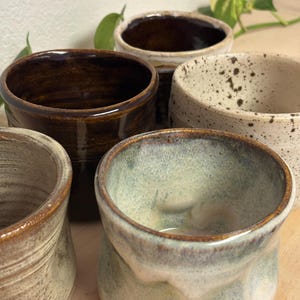 Puede incluir: Una colección de tazas de cerámica hechas a mano en varios colores y texturas. Las tazas presentan una gama de tonos tierra, incluyendo marrón, beige y blanco moteado. Las tazas tienen una estética rústica y orgánica.