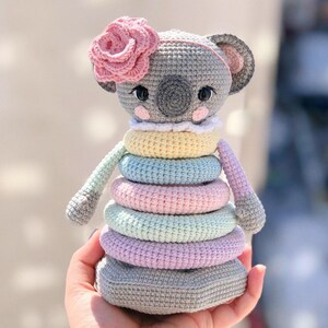 May include: A handmade crochet stacking toy shaped like a koala. The gray koala has a pink flower on its head and a stack of colorful rings in pastel shades. The toy is held in a person's hand.