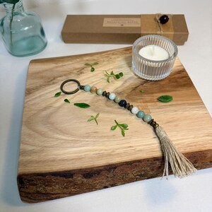 May include: A beaded keychain with a tassel, featuring a variety of earth-toned beads. The keychain is displayed on a wooden cutting board with a small candle in a glass holder. A small glass bottle and a box are also in the background.