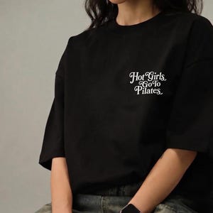 Può includere: T-shirt nero con la scritta bianca "Hot Girls Go To Pilates". La maglietta ha maniche corte e una vestibilità ampia. Il testo è scritto in corsivo ed è centrato sul petto.