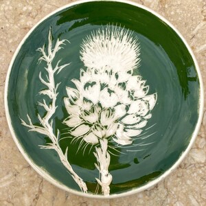 Puede incluir: Un plato redondo de cerámica con un esmalte verde oscuro. El plato presenta un diseño de cardo blanco en el centro. El borde del plato es de color crema claro. El plato es un objeto decorativo.