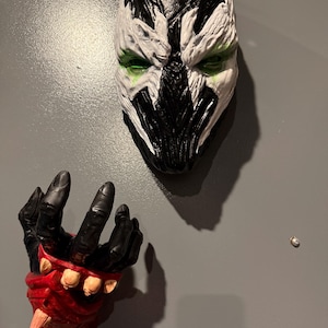 Puede incluir: Una máscara de pared con un diseño texturizado en blanco y negro con detalles verdes alrededor de las cuencas de los ojos, junto a una escultura de pared de una mano negra con un guantelete rojo y púas de color beige.