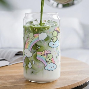 Puede incluir: Un vaso transparente en forma de lata lleno de una bebida verde, decorado con arcoíris, nubes y figuras de dibujos animados. La bebida se vierte de una jarra de cerámica. El vaso está sobre una superficie de madera.