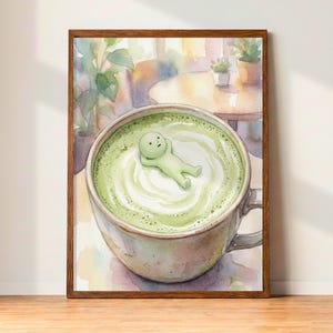 May include: Watercolor art print of a large cup of matcha latte with a whimsical green figure floating in the foam. The artwork is framed in a brown wooden frame and set against a blurred cafe background.