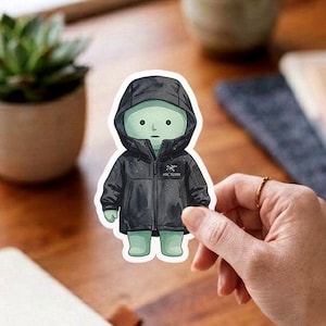 May include: A die-cut sticker featuring a pale green, cartoon-style character wearing a black hooded jacket. The jacket includes a visible white Arc'teryx logo on the chest area. The character has a simple, rounded design with a light green complexion.