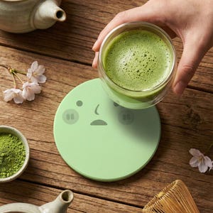 Puede incluir: Un vaso de té matcha verde espumoso se sostiene sobre un posavasos verde claro con una cara de dibujos animados. Un pequeño cuenco de polvo de matcha, un batidor de té y una tetera también están presentes.