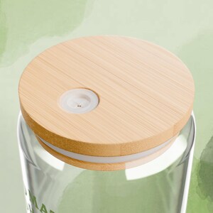 以下が含まれることがあります： 明るい茶色の竹の蓋が付いた透明なガラスタンブラー。蓋には白いプラスチック製のインサートと、縁に白いバンドが付いています。タンブラーには、薄い緑色の液体が部分的に入っています。背景は薄い緑色の水彩画です。