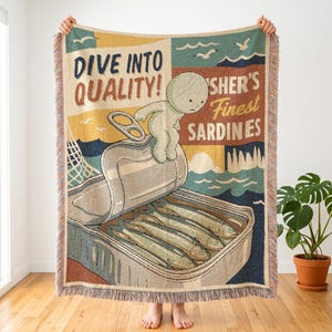 May include: A woven throw blanket featuring a vintage-style graphic of a sardine tin with the text DIVE INTO QUALITY and FISHER'S FINEST SARDINES. A cartoon character sits on the open lid above three sardines, with a sea and sky background.