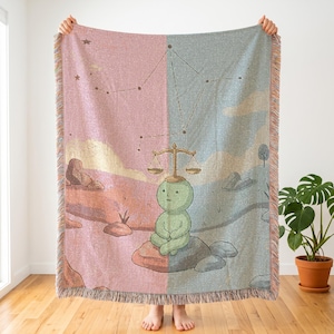 May include: A woven throw blanket featuring a split design with a pink side and a blue side. A green figure sits on a rock in the center, balancing a golden scale on its head. Above the figure, a constellation is depicted against the sky background.