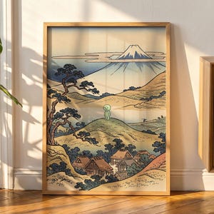 May include: A framed art print featuring a stylized landscape with a snow-capped mountain in the background, a small green character standing on a grassy hill in the center, and a village with thatched-roof houses nestled in the foreground.