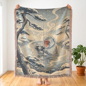 May include: A woven throw blanket featuring a detailed illustration of a Japanese macaque monkey submerged in a snowy hot spring while holding a camera. The scene includes stylized pine trees, swirling steam, and falling snow in shades of blue, white, and beige with a fringed edge.