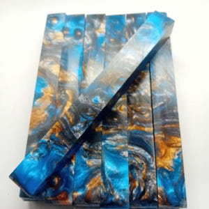 May include: A collection of rectangular resin blanks in shades of blue, gold, and grey. The resin has a marbled appearance, with swirls and patterns throughout. One blank is positioned diagonally across the others, showcasing the colour variations and textures.