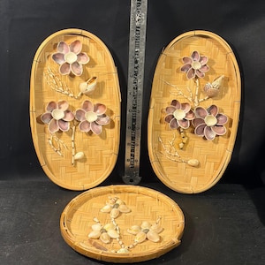 Peut inclure: Deux plateaux ovales et un plateau rond en bambou tressé, décorés de compositions florales faites de coquillages rose clair et blancs. Une règle en métal est placée entre les deux plateaux ovales, indiquant une longueur d'environ 30 cm.