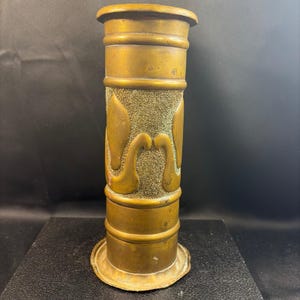 May include: A cylindrical brass vase featuring a wide, flat base and a flared rim. The exterior surface displays smooth horizontal bands at the top and bottom, while the central section is decorated with a textured finish and raised leaf motifs.