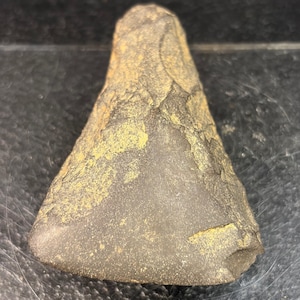 May include: A triangular stone tool with a dark gray, smooth, flat surface on one side and a rough, textured, brownish-gray surface on the other. The object features sharp, chipped edges along the sides and a pointed top with a wide, flat base.