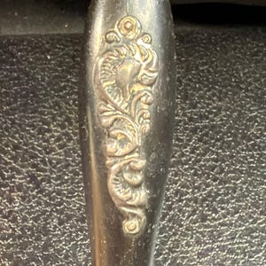 以下が含まれることがあります： 浮き彫りの花とスクロール模様が施されたダークメタリックなハンドル。表面は風化したような古びた風合いで、隆起した装飾部分には光沢があり、複雑なデザインの暗い凹部と明るく磨かれたディテールとのコントラストが際立っています。