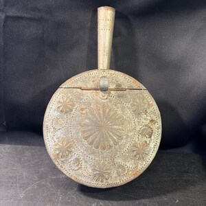 Op de afbeelding: Een ronde metalen haardblaasbalg met een lange, taps toelopende handgreep. Het oppervlak heeft een verweerde, zilverkleurige afwerking met reliëfbloemmotieven, stervormige patronen en gestippelde texturen op het voorpaneel.