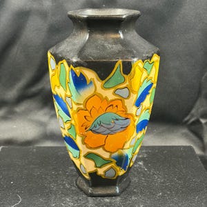 May include: A black vase with a hexagonal neck and base, featuring a vibrant, multi-coloured floral pattern. The design includes large orange flowers, blue leaves, and green accents set against a cream-coloured background, creating a bold contrast.