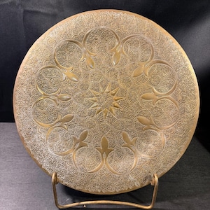 May include: A round brass plate featuring an intricate embossed pattern with a central star motif surrounded by repeating floral-like shapes and textured filigree details, supported by a simple metal stand for upright display.