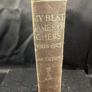 May include: A brown hardcover book spine with the text MY BEST GAMES OF CHESS 1908-1923 ALEKHINE printed in light-colored lettering. The spine features a small graphic of a chess queen piece near the bottom and has a textured fabric cover.