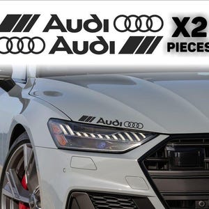 Può includere: Decalcomanie nere per auto Audi. Le decalcomanie includono il logo Audi e il testo "X2 PIECES". Le decalcomanie sono mostrate sul cofano di un'auto argentata.