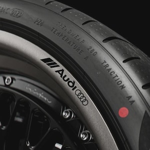 May include: Close-up of a black tire and wheel with the Audi logo. The tire has the text "TREADWEAR 280" and "TRACTION AA". The wheel is black with silver accents and bolts.