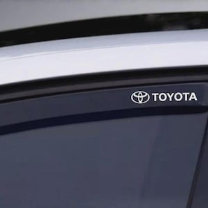 Może przedstawiać: Zbliżenie szyby samochodowej z białym logo i emblematem "TOYOTA". Logo znajduje się na czarnym daszku okiennym, z białym dachem samochodu widocznym w tle. Obraz jest dobrze oświetlony i skupia się na marce samochodu.