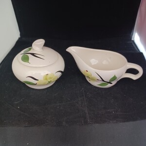 May include: Cream-colored ceramic sugar bowl with lid and creamer set. The set is decorated with yellow flowers, green leaves, and black stems. The sugar bowl has a rounded shape with a lid and a small handle. The creamer has a spout and a handle.