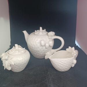 May include: A white ceramic tea set featuring a teapot, sugar bowl, and creamer. The teapot has a lid and handle, and the set is decorated with strawberry and floral details. The set is made of ceramic.