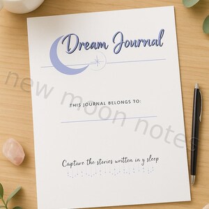 May include: A white Dream Journal with the text "Dream Journal" in blue script, a crescent moon graphic, and the phrase "Capture the stories written in y sleep." A black pen and a cup of coffee are on a wooden surface.