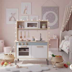 Könnte beinhalten: Eine Spielzeugküche aus Holz in Weiß und Blau, mit Mikrowelle, Herd, Spüle und Stauraum. Sie enthält Holzutensilien, Töpfe und Pfannen. Die Küche befindet sich in einem Kinderzimmer mit Bett, Kunstwerken und Spielzeugkörben.
