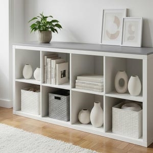 May include: A white cube storage unit with a gray top, featuring eight open compartments. The shelves display books, vases, and decorative baskets. A potted plant and framed art pieces sit on top. The unit is placed on a wooden floor.